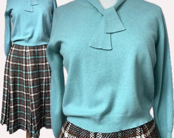 May include: A light blue long-sleeve jumper with a tie-neck detail paired with a brown, white, and light blue plaid pleated skirt. The jumper features a solid knit texture, while the skirt displays a repeating checkered pattern.