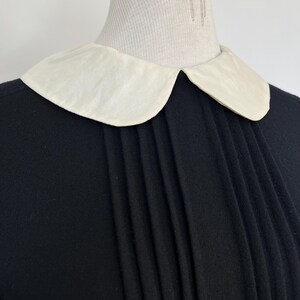 May include: A black dress with a white Peter Pan collar and pleated detail at the front.