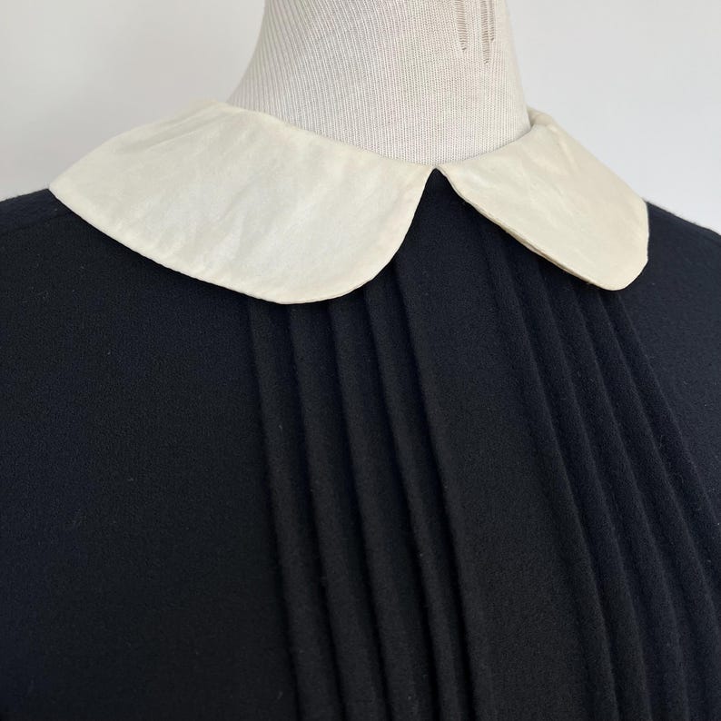 May include: A black dress with a white Peter Pan collar and pleated detail at the front.
