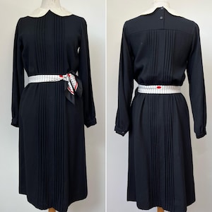 May include: A black midi dress with a white Peter Pan collar and long sleeves. The dress has a pleated skirt and is cinched at the waist with a white belt with black polka dots. A black and white polka dot scarf is tied around the waist.
