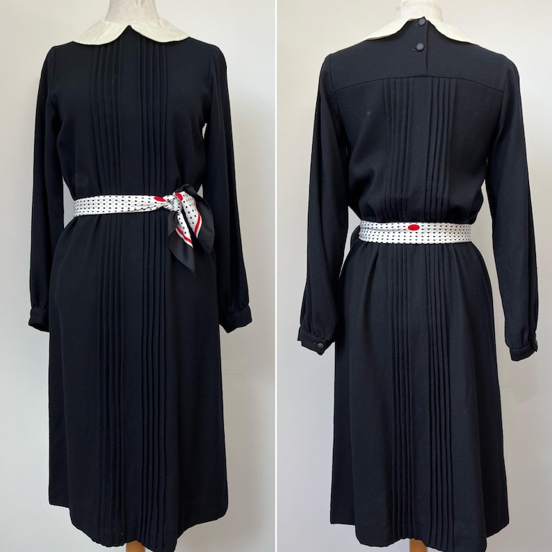 May include: A black midi dress with a white Peter Pan collar and long sleeves. The dress has a pleated skirt and is cinched at the waist with a white belt with black polka dots. A black and white polka dot scarf is tied around the waist.