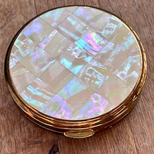 May include: A round compact mirror with a gold-toned metal frame and a lid featuring a grid of iridescent, square mother-of-pearl tiles. The surface of the tiles displays shifting hues of white, pink, and blue, while a small gold latch is visible.