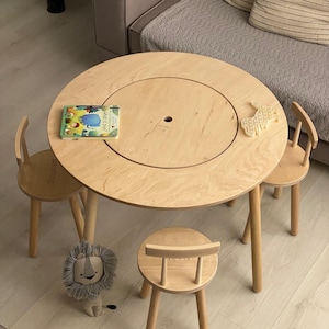 Op de afbeelding: Een ronde tafel van lichtgekleurd hout met een centraal rond inzetstuk en drie bijpassende houten krukjes. Een klein boekje getiteld What is poo? en een lichtgekleurd diervormig speeltje liggen op het tafelblad. Een pluche leeuw staat op de vloer.