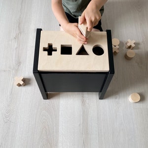 May include: A black wooden toy box with a light wood top. The top has cutouts in the shape of a cross, square, triangle, and circle. A child is playing with wooden blocks that fit into the cutouts.