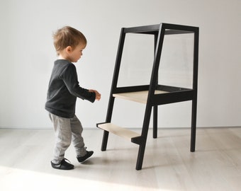 May include: A black wooden learning tower with a tall frame and two light-colored wooden steps. The structure features a high safety rail at the top and a sturdy base, designed to provide elevation for reaching countertops or tables safely.
