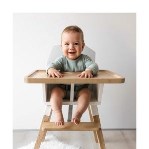 Op de afbeelding: Een lachende baby zit in een houten kinderstoel met een licht dienblad en witte accenten. De kinderstoel heeft een natuurlijke houten afwerking en staat op een zacht wit tapijt. De baby draagt een lichtgroen shirt met lange mouwen.