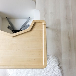 Op de afbeelding: Een moderne, witte kinderstoel met een natuurhouten blad en grijze veiligheidsriemen. De kinderstoel staat tegen een lichte houten vloer en een pluizig wit tapijt.