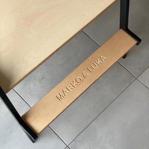 May include: A wooden learning tower with a light-colored plywood platform and a black frame. The lower step features the engraved text MARKO & LUKA in capital letters. The structure is designed with a sturdy base and a smooth, natural wood finish.