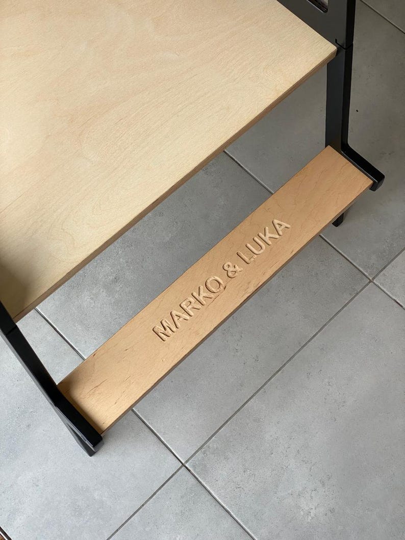 May include: A wooden learning tower with a light-colored plywood platform and a black frame. The lower step features the engraved text MARKO & LUKA in capital letters. The structure is designed with a sturdy base and a smooth, natural wood finish.