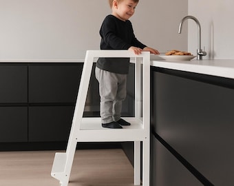 May include: A white wooden learning tower with a single step and a safety railing, designed to elevate a child to counter height. The structure features a sturdy frame with four legs and a flat platform for standing, finished in a smooth white paint.