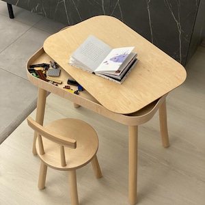Op de afbeelding: Een tafel- en stoelset van lichtgekleurd hout. De tafel heeft een afneembaar blad dat een opbergvak onthult met kleine plastic bouwstenen. Een open boek met witte pagina's en illustraties ligt op het oppervlak van de tafel.