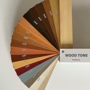 May include: A fan-shaped wood tone color swatch set featuring various shades of brown, tan, red, blue, and white. Each wooden sample is labeled with a numerical code. A white rectangular handle is attached, labeled with the text WOOD TONE PIDkids.