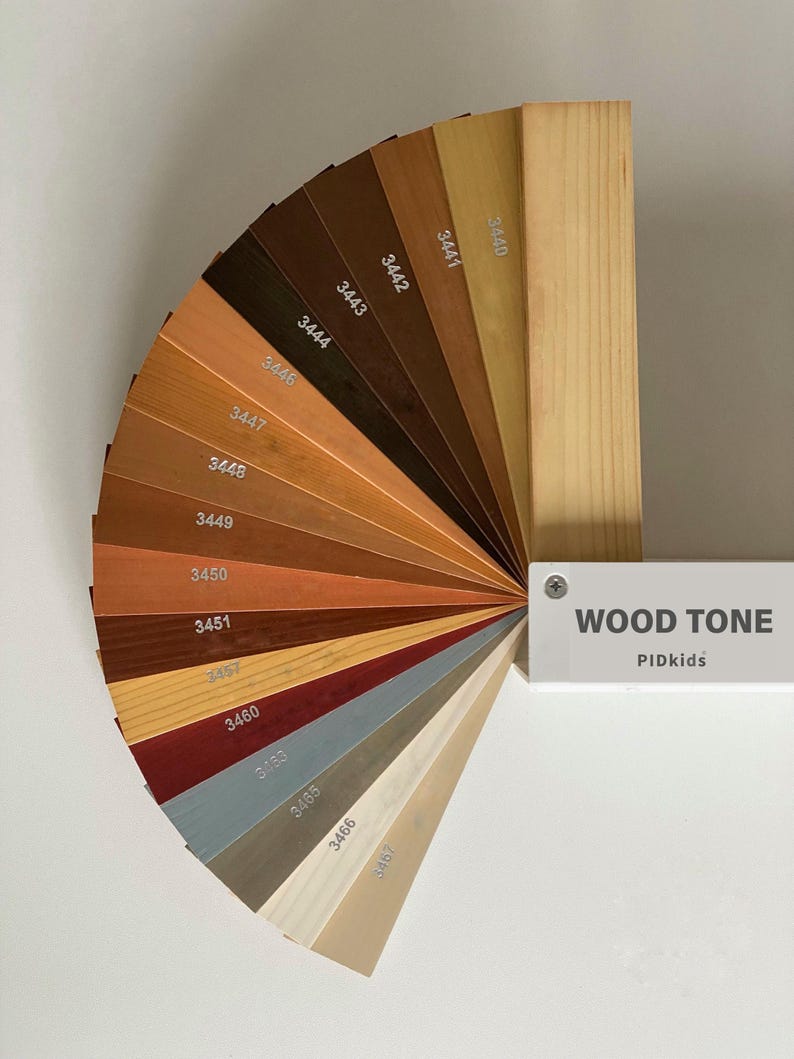 May include: A fan-shaped wood tone color swatch set featuring various shades of brown, tan, red, blue, and white. Each wooden sample is labeled with a numerical code. A white rectangular handle is attached, labeled with the text WOOD TONE PIDkids.
