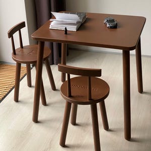 May include: A brown wooden table with four rounded legs and two matching round stools with backrests. The table surface holds an open white book and a small black toy car. Each stool features a small engraved word on the seat surface.