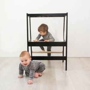 May include: A black wooden learning tower with a clear acrylic panel and a natural wood step. The structure features a rectangular frame design with a platform for standing and a lower step, providing a stable elevated area for children to reach surfaces.