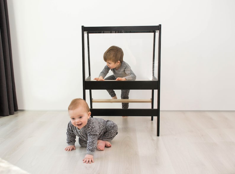May include: A black wooden learning tower with a clear acrylic panel and a natural wood step. The structure features a rectangular frame design with a platform for standing and a lower step, providing a stable elevated area for children to reach surfaces.