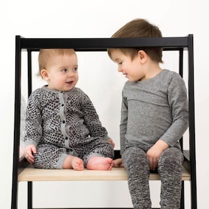 May include: A black wooden learning tower with a light wood platform. One child wears a gray leopard print long-sleeve romper with a button-down front. The other child wears a plain gray long-sleeve shirt and dark gray heathered leggings.