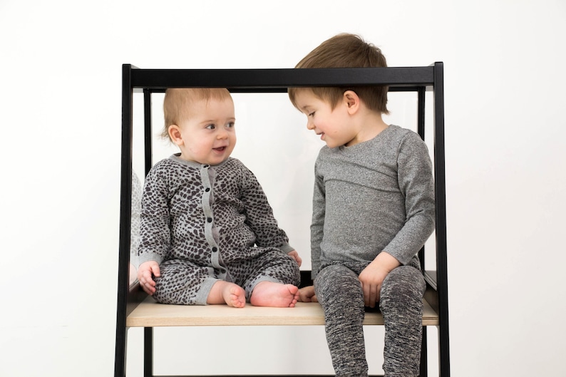 May include: A black wooden learning tower with a light wood platform. One child wears a gray leopard print long-sleeve romper with a button-down front. The other child wears a plain gray long-sleeve shirt and dark gray heathered leggings.