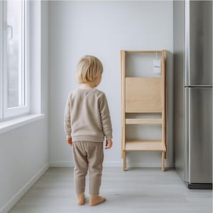 Könnte beinhalten: Ein Tritthocker aus Naturholz mit Falt-Design. Der Hocker hat zwei Stufen und eine Rückenlehne. Ein Kind in einem beigefarbenen Trainingsanzug steht in einem Raum mit Fenster und Kühlschrank in der Nähe des Hockers.
