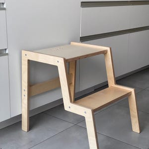 May include: A light-coloured wooden step stool with a two-tiered design. The stool has a rectangular top and a lower step, supported by angled legs. The wood grain is visible, and the stool is set against a neutral background.