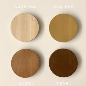 May include: Four circular wooden discs arranged in a grid, each displaying a different wood stain color. The labels read Natural, Old Oak, Hazel, and Teak. Each disc shows a distinct wood grain pattern, ranging from light beige to dark brown tones.