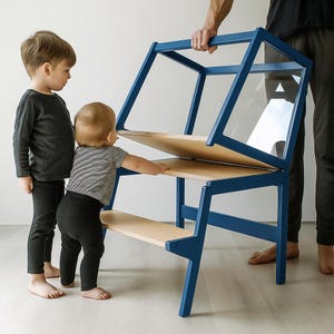 May include: A blue wooden learning tower with a light wood finish on the steps and platform. The structure features a frame with a clear acrylic panel on the upper section, designed to provide a stable elevated standing area for children.