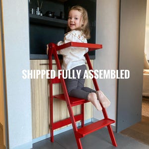 May include: A vibrant red wooden kitchen step stool with a small platform and two steps. The stool is designed to help children reach countertops. The text "SHIPPED FULLY ASSEMBLED" is displayed in white.