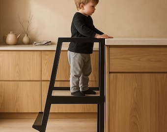 May include: A black wooden learning tower with a rectangular frame and a single step, designed to provide elevation. The structure features a sturdy base and a safety rail, allowing for stable positioning near a kitchen counter surface.