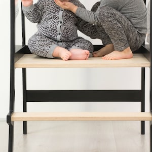 May include: A black learning tower with a light wood platform and a lower step. The structure features a tall frame with vertical supports and a horizontal crossbar, providing a stable elevated standing area for children to reach countertops.
