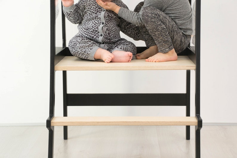 May include: A black learning tower with a light wood platform and a lower step. The structure features a tall frame with vertical supports and a horizontal crossbar, providing a stable elevated standing area for children to reach countertops.