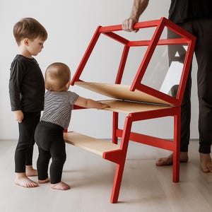 May include: A red wooden learning tower with a foldable design, featuring light-colored wooden steps and a platform. The structure includes a hinged upper frame with clear panels, allowing it to convert into a desk or table for children.