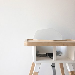 Op de afbeelding: Een moderne kinderstoel met een wit frame en accenten van natuurlijk hout. De stoel heeft een dienblad, een rugleuning en een grijze veiligheidsgordel. De poten zijn schuin en hebben een tweekleurig ontwerp. De stoel is ontworpen voor een baby of peuter.