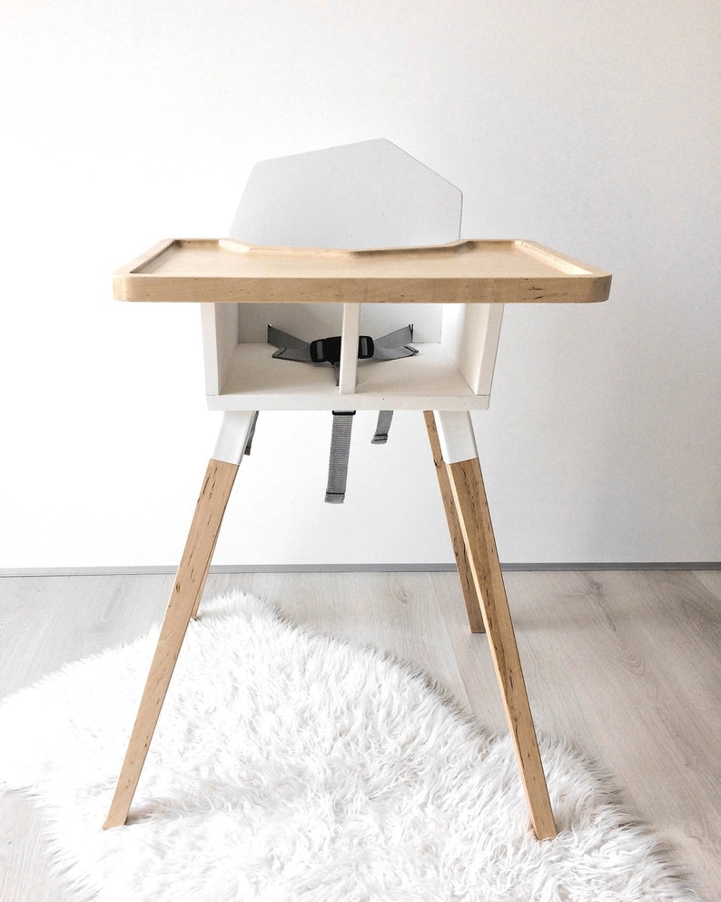 Op de afbeelding: Een moderne kinderstoel met een dienblad en poten van natuurlijk hout, een witte zitting en een grijze veiligheidsgordel. De stoel heeft een minimalistisch ontwerp en is geschikt voor een baby of peuter.