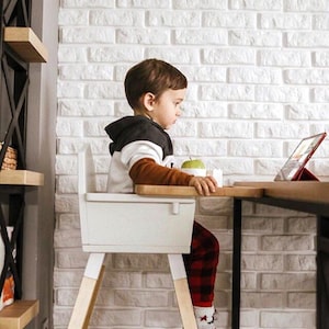 Op de afbeelding: Een kind zit in een witte kinderstoel met houten poten en kijkt naar een tablet op een houten tafel. Het kind draagt een zwart-witte hoodie, een bruin shirt met lange mouwen en een rode geruite broek. Er ligt een appel op tafel.