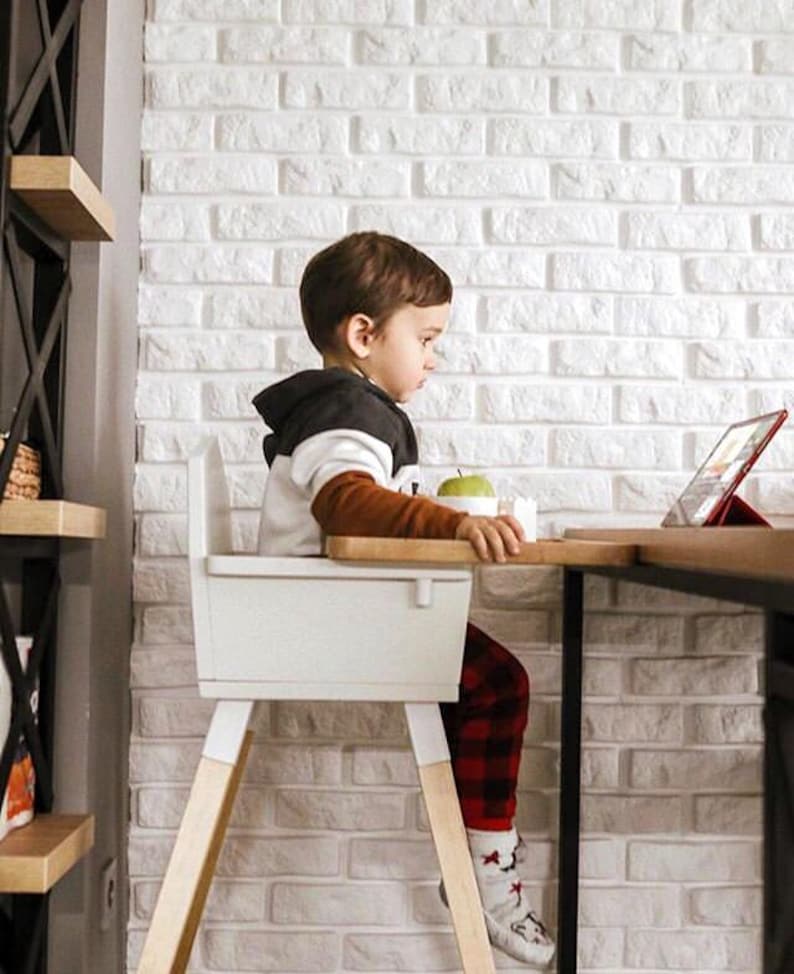 Op de afbeelding: Een kind zit in een witte kinderstoel met houten poten en kijkt naar een tablet op een houten tafel. Het kind draagt een zwart-witte hoodie, een bruin shirt met lange mouwen en een rode geruite broek. Er ligt een appel op tafel.