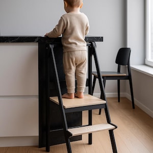 May include: A black and natural wood step stool with two steps. A child is standing on the top step, facing away from the camera. The step stool is next to a black countertop and a black chair.