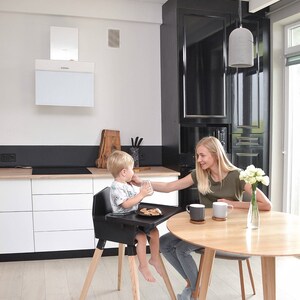 Op de afbeelding: Een zwarte kinderstoel met houten poten. Een kind zit in de stoel aan een tafel. Een vrouw zit aan een ronde houten tafel met twee mokken en een vaas met bloemen. De keuken heeft witte kasten en een zwart aanrecht.