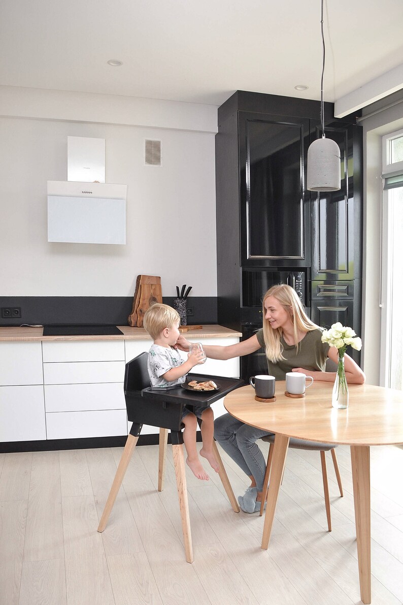 Op de afbeelding: Een zwarte kinderstoel met houten poten. Een kind zit in de stoel aan een tafel. Een vrouw zit aan een ronde houten tafel met twee mokken en een vaas met bloemen. De keuken heeft witte kasten en een zwart aanrecht.