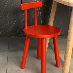 May include: A small, pillar-box red wooden chair with a round seat and a rectangular backrest. The chair has four cylindrical legs and a simple design. The chair is positioned in front of a dark grey marble-patterned wall.