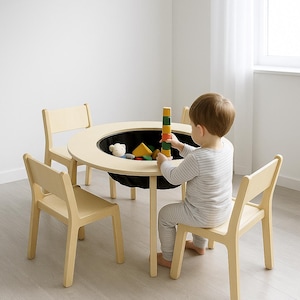 May include: A light wood-toned children's play table with a black fabric storage bin in the center, surrounded by three matching chairs. A child is playing with colorful wooden blocks. The table is round and has a diameter of approximately 30 inches.