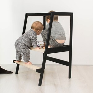 May include: A black wooden learning tower with two light-colored wooden steps. The structure features a tall, open-frame design with a safety rail at the top. The tower is positioned on a light-colored floor, providing a stable platform for children.