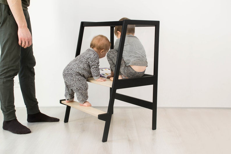 May include: A black wooden learning tower with two light-colored wooden steps. The structure features a tall, open-frame design with a safety rail at the top. The tower is positioned on a light-colored floor, providing a stable platform for children.