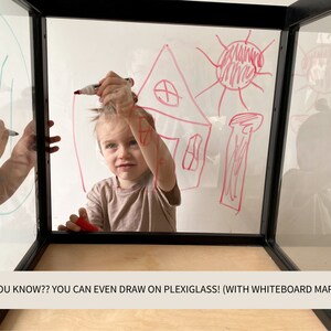 May include: A clear plexiglass cube with a black frame and a light wood base. The transparent panels feature red and green marker drawings of a house and sun. The text on the image reads: DID YOU KNOW?? YOU CAN EVEN DRAW ON PLEXIGLASS! (WITH WHITEBOARD MARKERS)