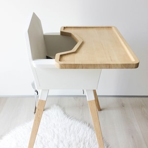 Op de afbeelding: Een moderne witte kinderstoel met een blad en poten van natuurlijk hout. De stoel heeft een ingebouwd veiligheidsharnas en een minimalistisch ontwerp. De blad is ongeveer 46 cm breed.