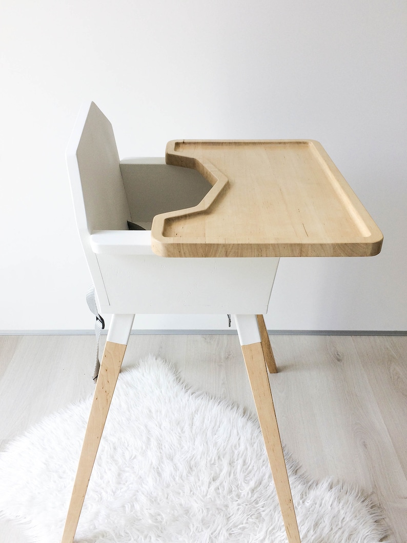 Op de afbeelding: Een moderne witte kinderstoel met een blad en poten van natuurlijk hout. De stoel heeft een ingebouwd veiligheidsharnas en een minimalistisch ontwerp. De blad is ongeveer 46 cm breed.