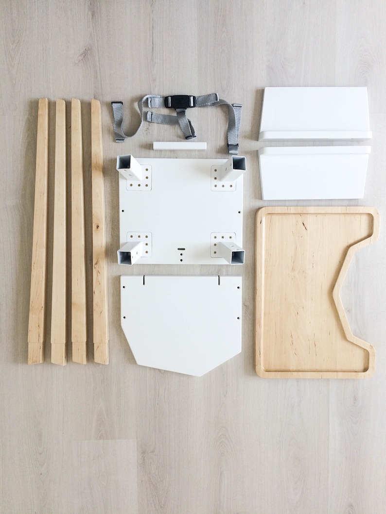 Op de afbeelding: Een bovenaanzicht toont de gedemonteerde onderdelen van een kinderstoel. De onderdelen omvatten houten poten, witte zittingonderdelen, een houten dienblad en een grijze veiligheidsgordel. De stukken zijn gerangschikt op een licht houten oppervlak.