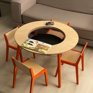 Puede incluir: Una mesa de actividades redonda de madera con patas de color naranja brillante y un compartimento central de malla, acompañada de tres sillas a juego. Un pequeño coche de juguete gris y un libro infantil abierto con dibujos de obreros.