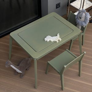May include: A sage green rectangular table with two matching chairs. A white dinosaur-shaped silicone sensory toy rests on the tabletop. A gray plush dinosaur toy sits on the floor nearby, and a gray plush lion toy rests on one of the chairs.