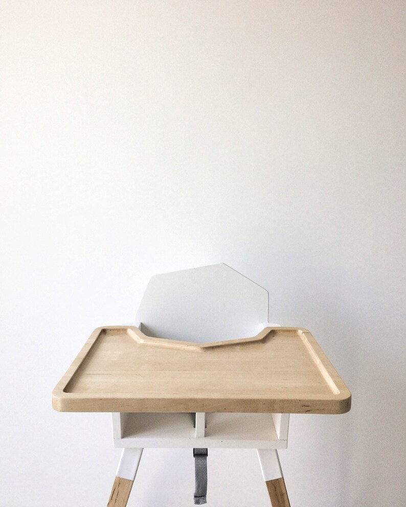 Op de afbeelding: Een moderne kinderstoel met een licht houten blad en witte accenten. De stoel heeft een witte rugleuning en een veiligheidsriem. De poten zijn wit met voeten in natuurlijk hout. De lade is ongeveer 51 cm breed.