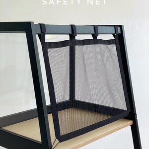 May include: A black mesh safety net attached to a black wooden frame with light wood shelving. The text SAFETY NET is printed in white above the mesh. The net features black fabric borders and is secured with straps to the top horizontal bar.